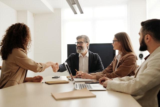 A recruitment team of 3 people sat around a table. A bearded man shakes hands with an interviewee who has their back to the camera. Why Employers Should Recruit Apprentices: The Economic Case