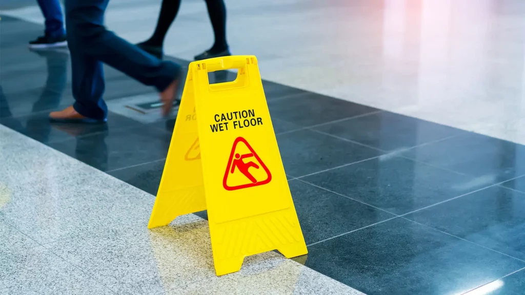 Health and safety : Image of a yellow floor sign that says "caution wet floor" on a tiled floor with people walking in the background. 