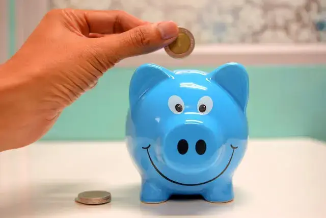 Image of a hand putting a coin into a blue piggy bank with a smiling face. The changes to apprenticeship funding could save businesses money.